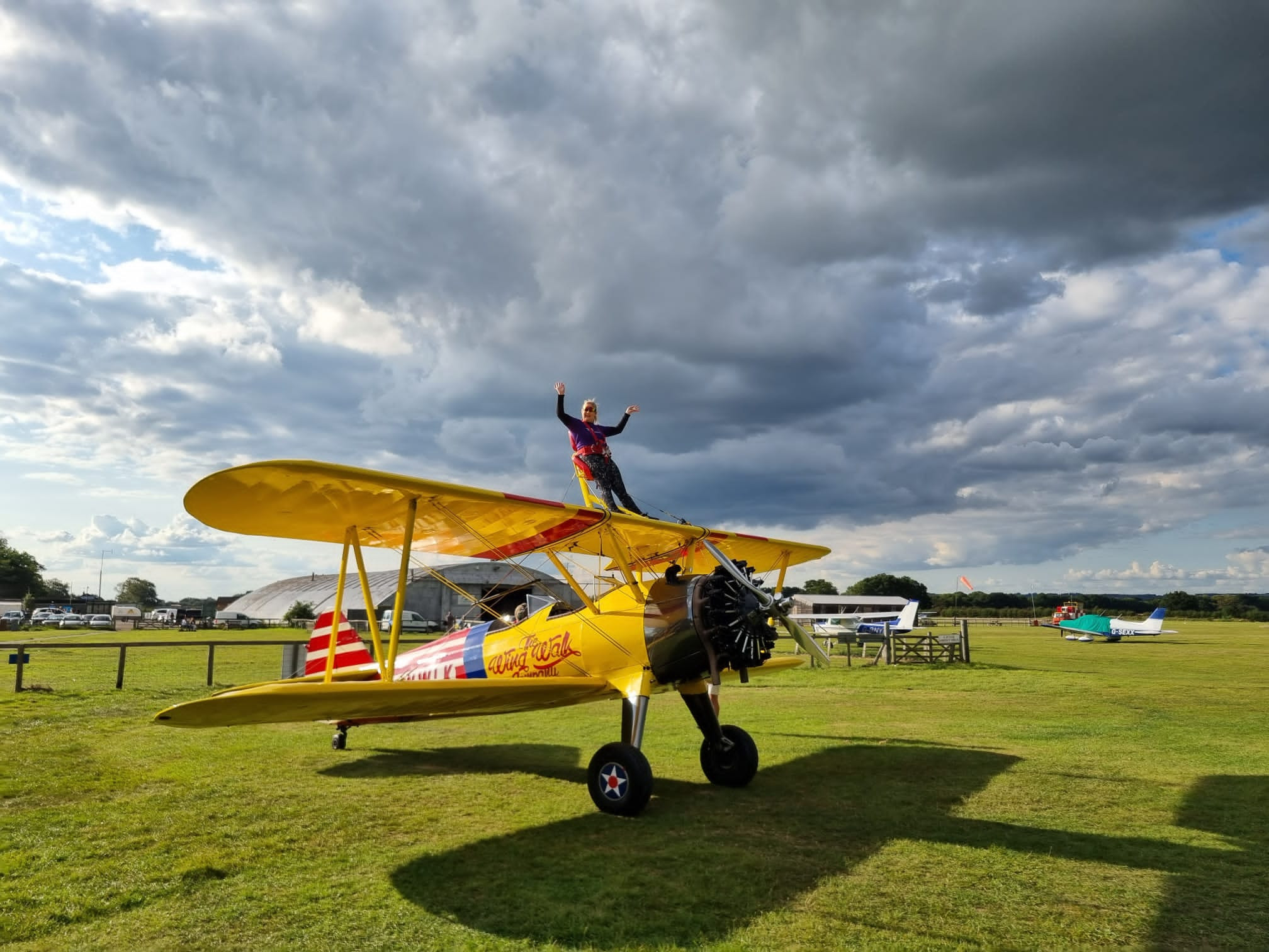Carly Larkin ready for her flight - she is standing on the top wing of a yellow bi-plane which is parked in a field.