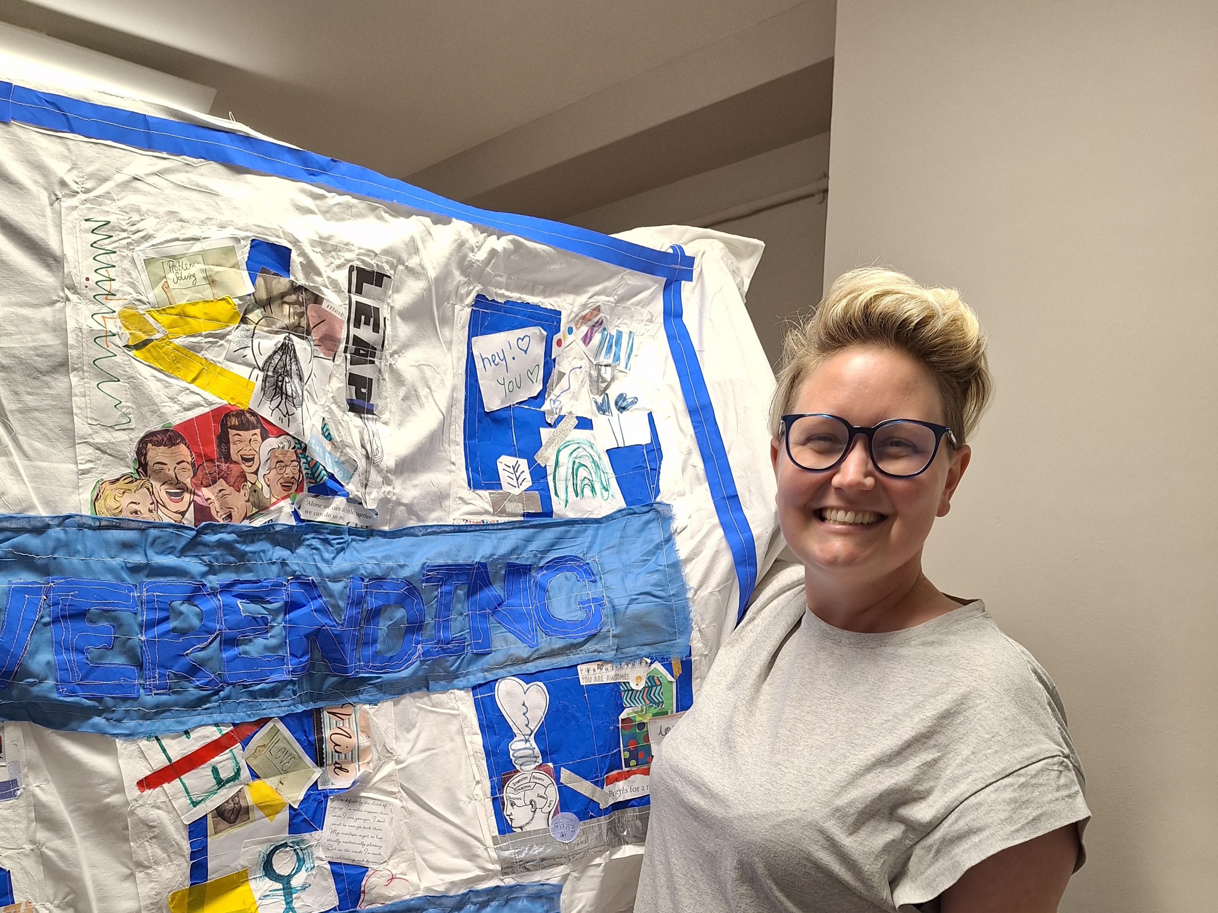 Emma Pope with her artwork, Hanging by a Thread. She is holding it up and standing next to it - it is a bed sheet with embroidery on