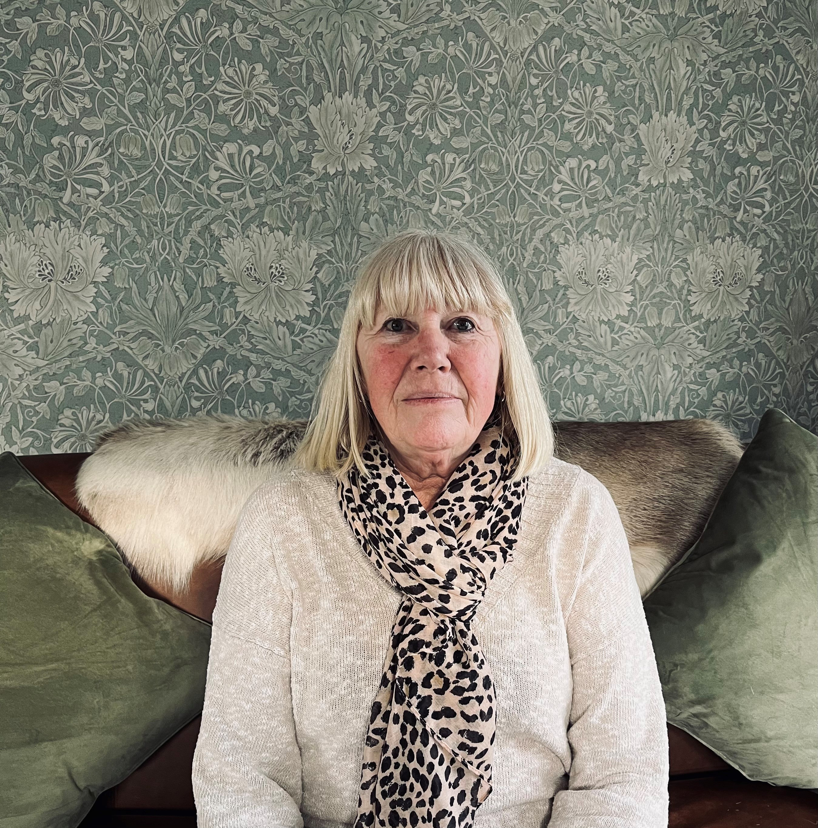 Jayne Stokes, pictured on a sofa with cushions and green wallpaper behind