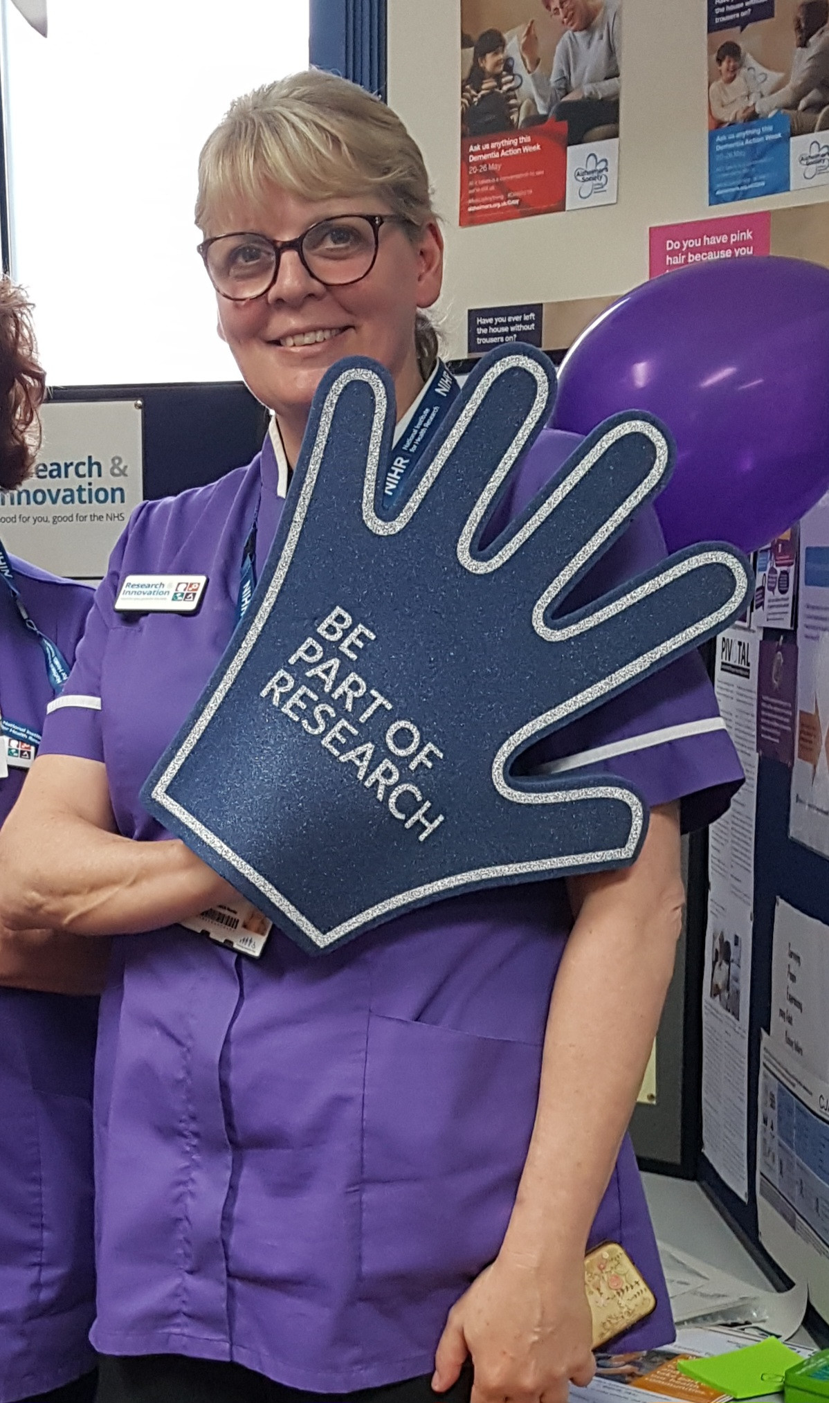 Lead research nurse Ruth Hulbert, who is retiring. She is wearing a purple tunic and holding a giant hand that says Be Part of Research