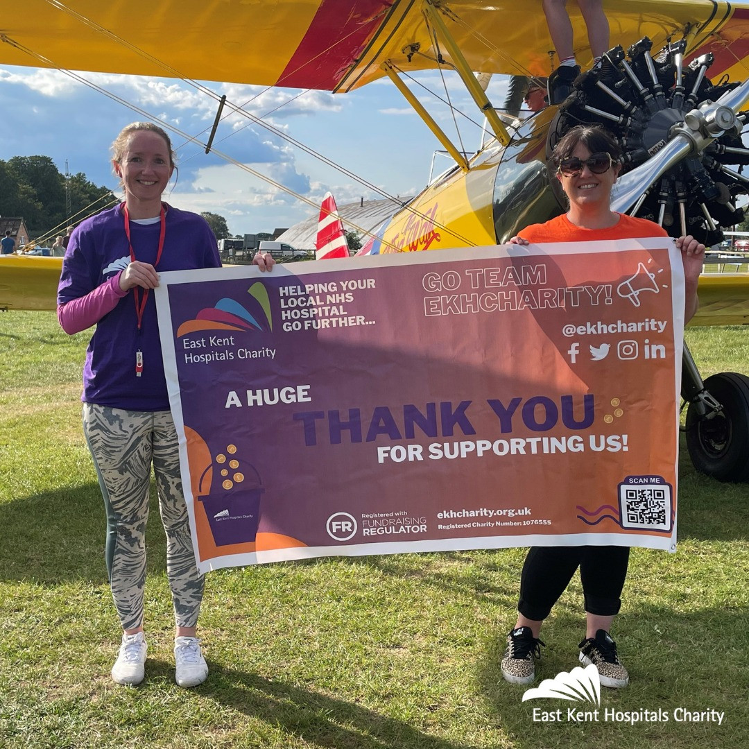 Louise Linden with Dee Neligan from East Kent Hospitals Charity. They are standing in front of a yellow biplane holding a banner advertising East Kent Hospitals Charity