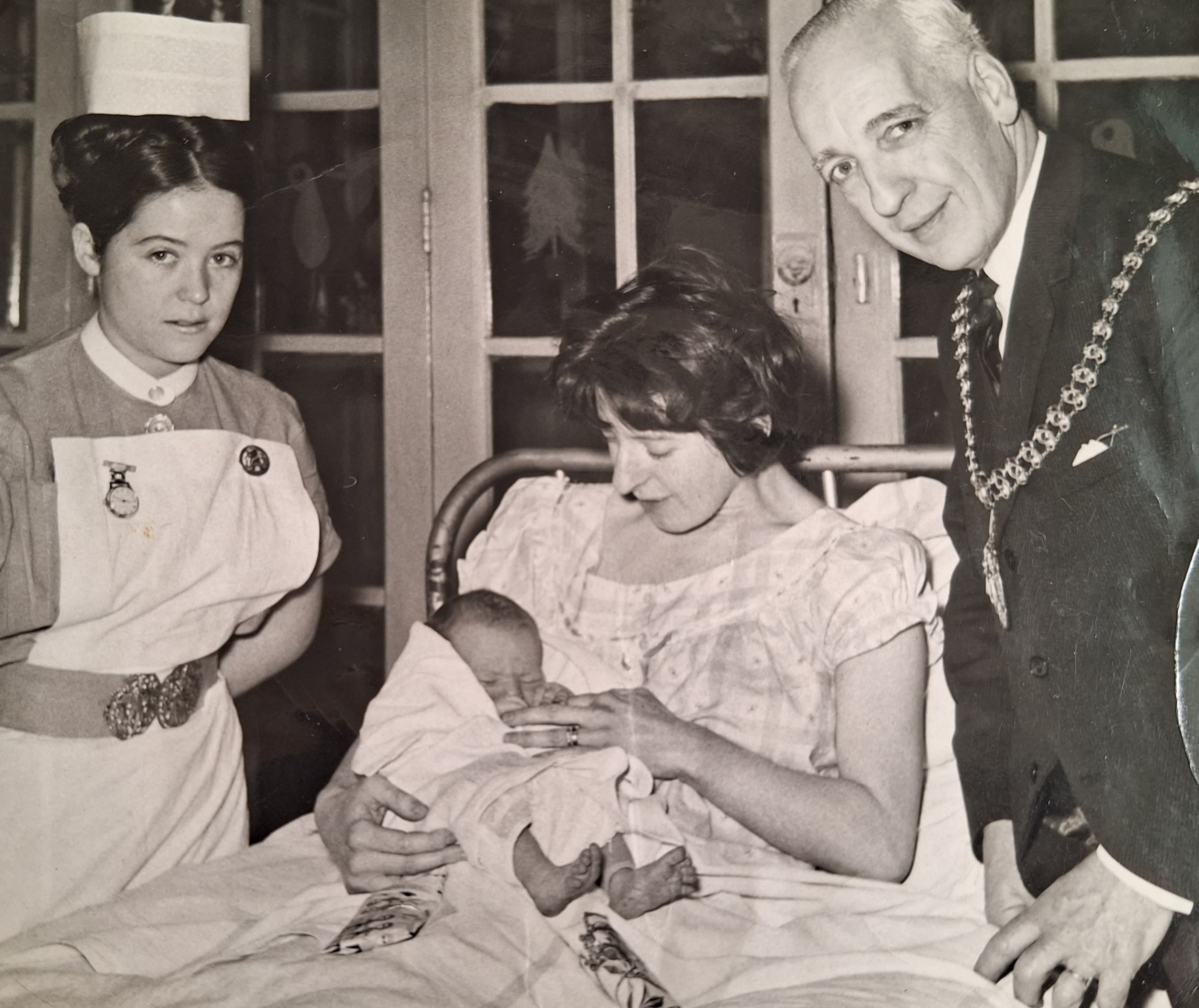 Marcella Warburton as a young nurse, during a civic visit to a Christmas day baby. Image shows Marcella in old-fashioned nurse's uniform, with a woman in bed holding a baby and a man wearing mayoral chains