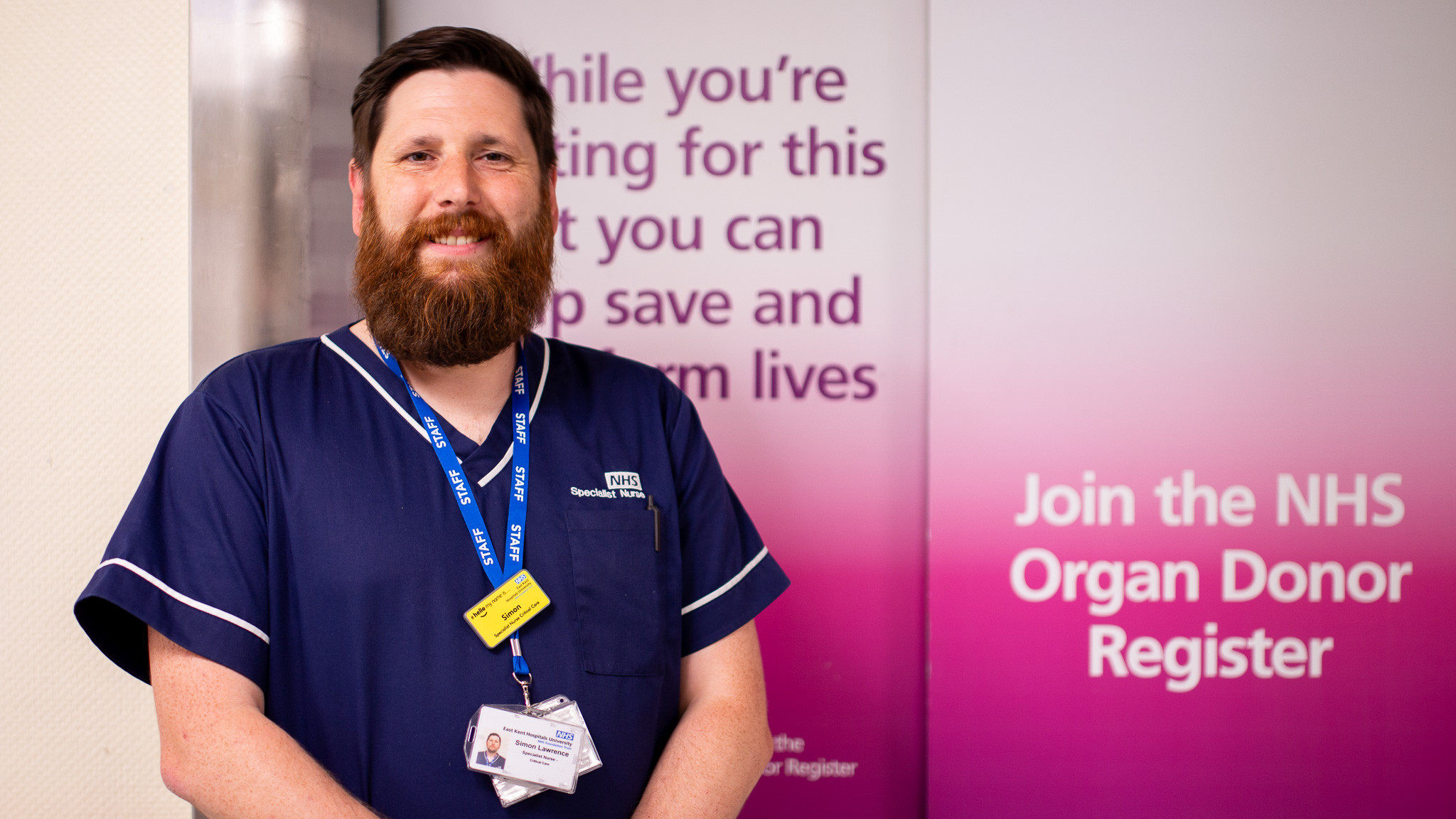 Simon Lawrence, specialist organ donation nurse. He is wearing a blue uniform and standing in front of a sign advertising the NHS organ donor register