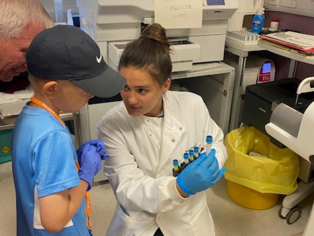 Ollie Bagiel is shown a sample in the lab. Image shows Ollie in a blue football kit and hat, being shown the sample by a woman in a lab coat with machines in the background