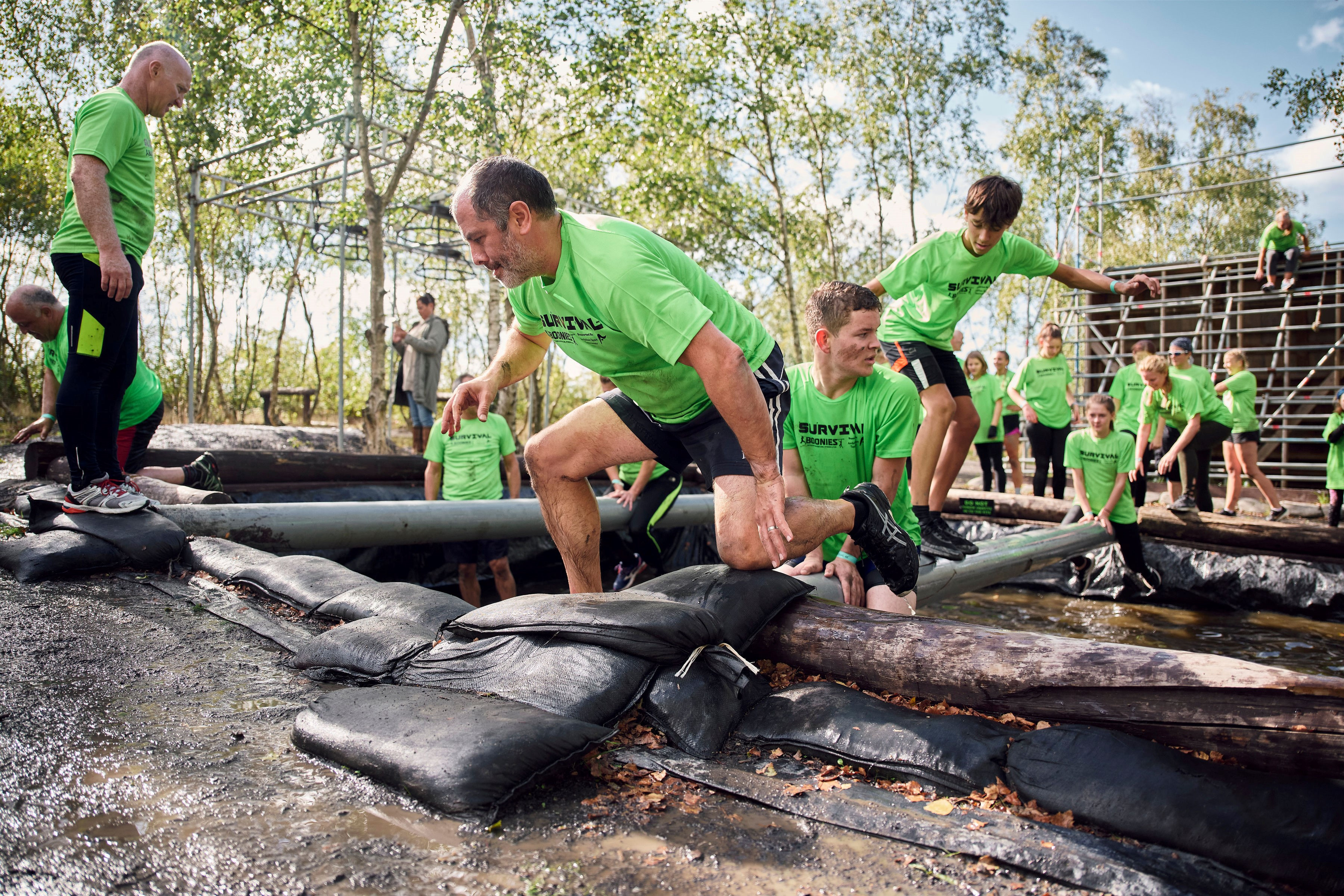 People take part in the Survival challenge at Betteshanger park. Image shows several people in green t-shirts tackling an obstacle that involves a log over a muddy put
