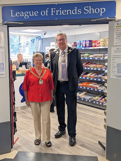 The Lord Mayor of Canterbury, Cllr Jean Butcher, and Sheriff Cllr Tom Mellish, at the League of Friends shop. They are both wearing civic chains and standing in the doorway of the shop with the sign above them