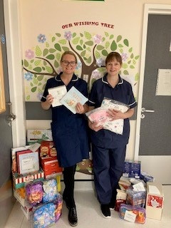 Vicky Vining and colleague Louise Sanders with some of the donations. They are wearing dark blue uniforms and standing in front of a tree mural on a wall, holding packages with others at their feet.