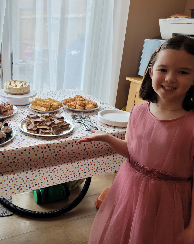 Sophie with some of the treats she made for the cake sale. Image shows a young girl in a pink dress standing next to a table with a spotty tablecloth and various sweet treats on plates