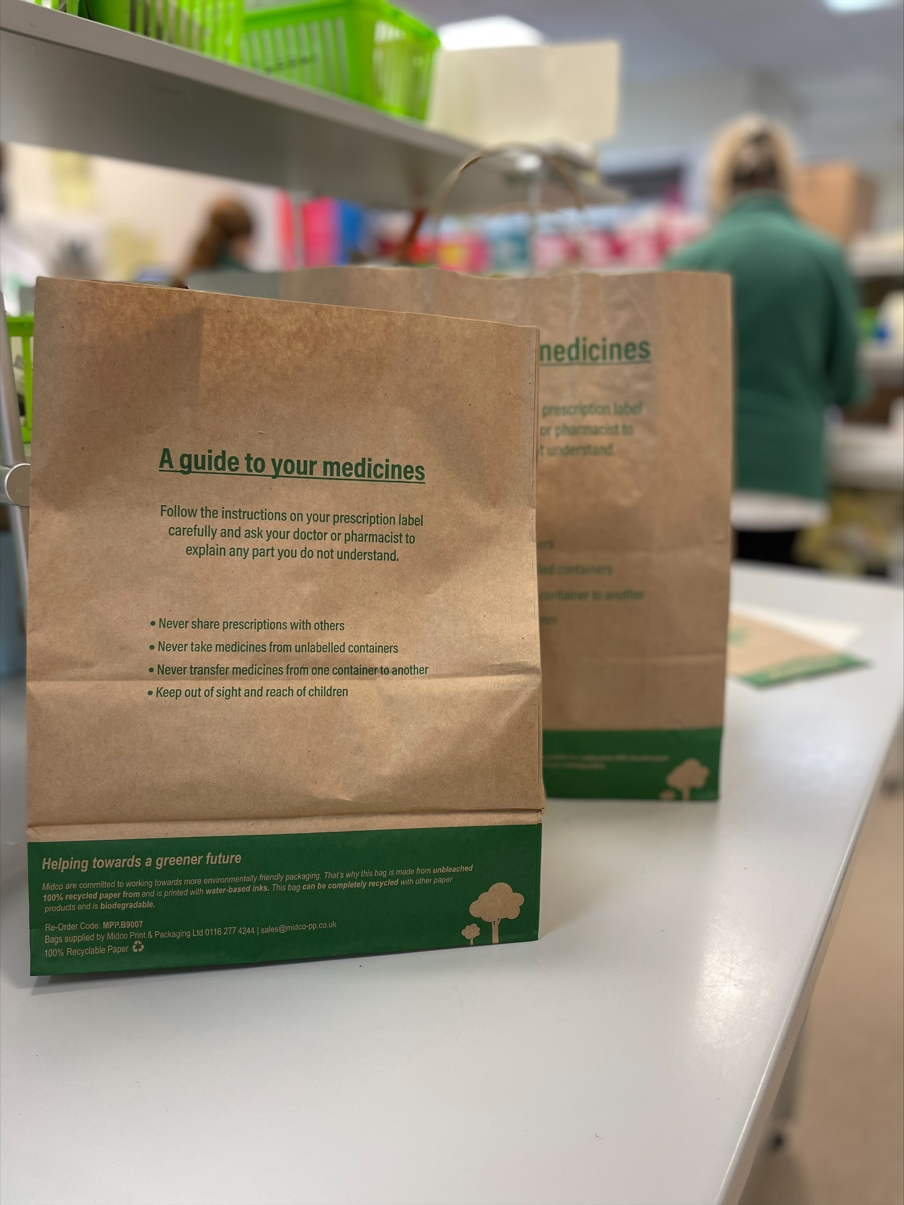 Paper bags, used in pharmacy instead of plastic bags. Image shows two brown paper bags with green writing on, on the counter in the pharmacy department. Staff can be seen in the background working.